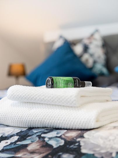 On a bed lie two folded white towels. On top, there is a bottle with green contents and a decorative pillow in the background.
