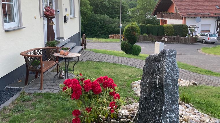 Eine gemütliche Hausecke mit einem Blumengarten und einem dekorativen Stein. Im Hintergrund sind Hügel und ein klarer Himmel zu sehen.