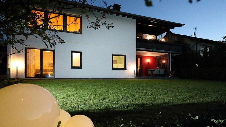 Ein modernes Haus mit großen Fenstern, beleuchtet bei Nacht. Der Garten ist grün und hat dekorative Beleuchtungselemente.