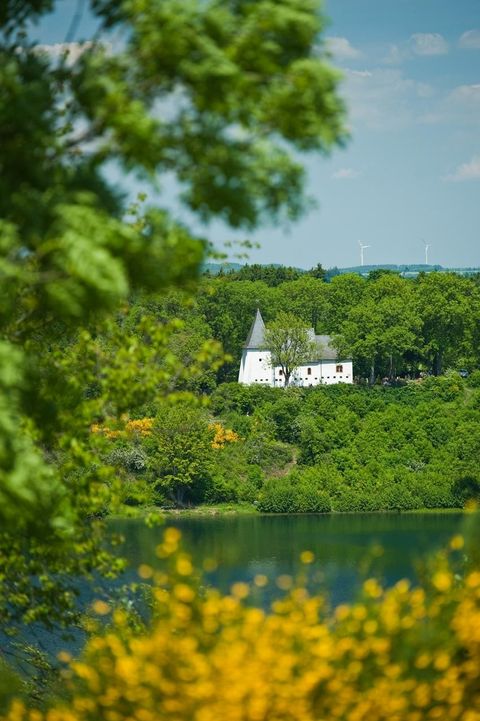 Ein weißes Gebäude mit Türmen liegt malerisch an einem See, umgeben von grüner Natur. Die Szenerie strahlt Ruhe und Idylle aus.
