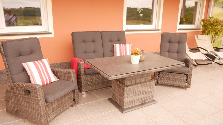 Eine gemütliche Terrasse mit rattanartigen Möbeln und bequemen Kissen. Der Tisch steht in der Mitte und ein Blumentopf sorgt für eine einladende Atmosphäre.