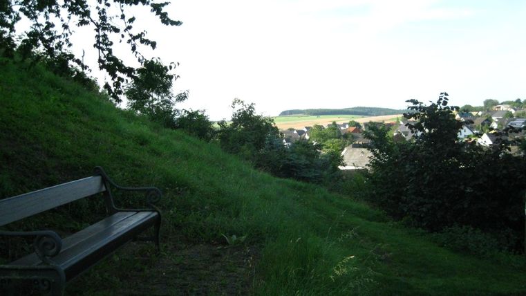 Eine ruhige Landschaft mit einer Bank im Vordergrund und grünen Wiesen im Hintergrund. Im Hintergrund sind einige Häuser und ein sanfter Hügel zu sehen.