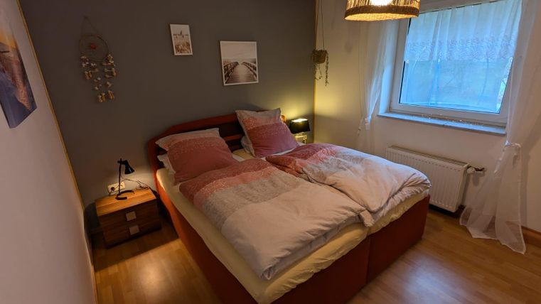 Ein gemütliches Schlafzimmer mit einem großen Doppelbett und sanften Farben. Ein Fenster sorgt für Tageslicht und eine entspannte Atmosphäre.