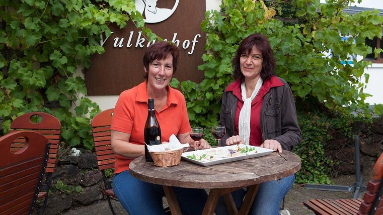 Zwei Frauen sitzen an einem Tisch im Freien und genießen ein Getränk. Im Hintergrund ist ein Schild mit dem Namen „Vulkanhof“ und grüne Weinreben zu sehen.