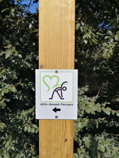 Ein Schild mit dem Hinweis auf den Aktiv-Gesund-Parcours. Es zeigt ein Symbol für Bewegung und Gesundheit.