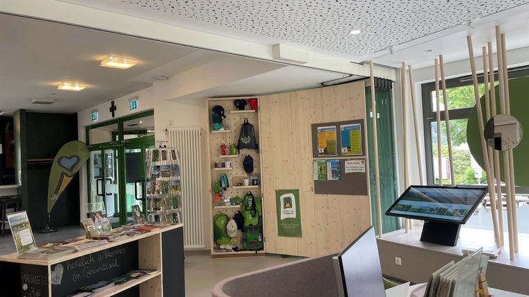 Ein moderner Empfangsbereich mit einem Informationsschalter und Ausstellungsregalen. Im Hintergrund sind digitale Informationshilfen und eine grüne Wand sichtbar.