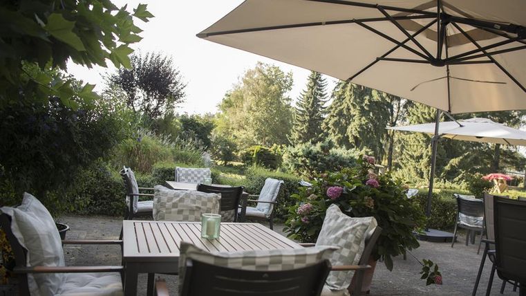 Ein gemütlicher Gartensitzbereich mit Tischen und Stühlen unter Sonnenschirmen. Umgeben von einer grünen Landschaft und blühenden Pflanzen.