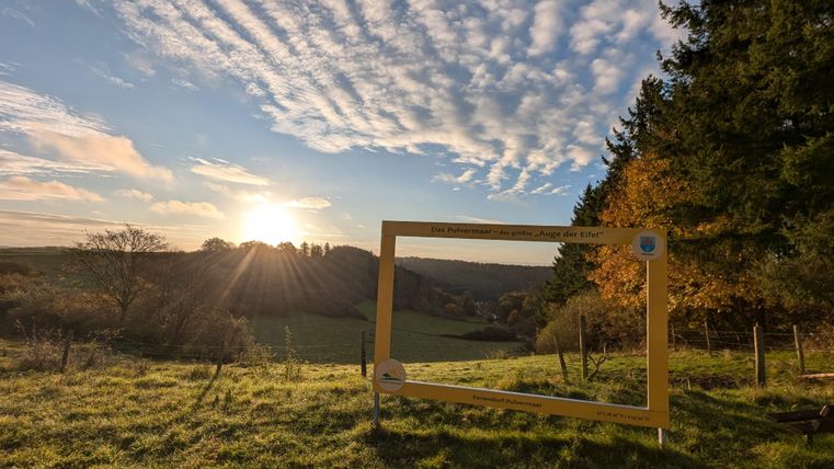 Ein malerischer Ausblick auf eine grüne Landschaft mit einem Sonnenuntergang im Hintergrund. Im Vordergrund steht ein gelber Rahmen, der als Fotopunkt dient.