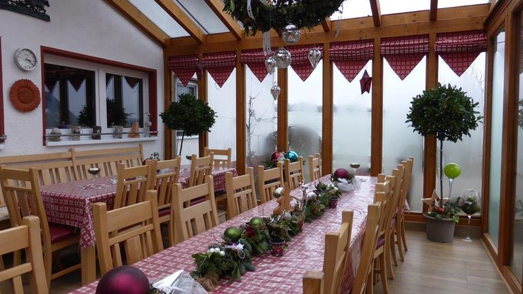Ein heller Wintergarten mit langen Tischen und Stühlen aus Holz. Die Tische sind festlich mit Weihnachtsdekorationen geschmückt.