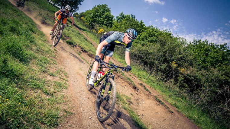 Drei Mountainbiker fahren einen staubigen Pfad in der Natur. Es ist ein sonniger Tag mit grünem Gras und Bäumen im Hintergrund.