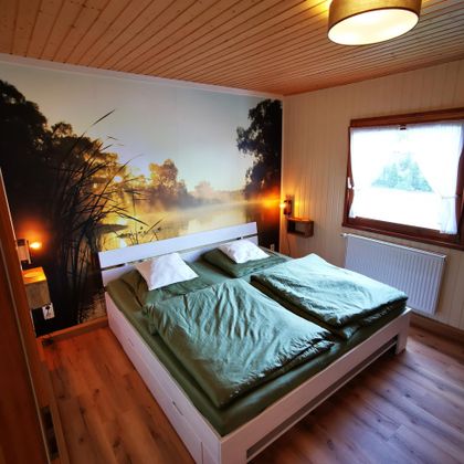 Ein gemütliches Schlafzimmer mit einem großen Bett und grüner Bettwäsche. An der Wand ist eine malerische Landschaft mit einem See und Bäumen zu sehen.