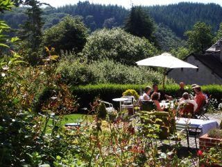 Ein sonniger Garten mit gemütlichen Sitzplätzen und einer Gruppe von Menschen, die sich unterhalten. Die Umgebung ist grün und einladend, umgeben von Bäumen und Blumen.