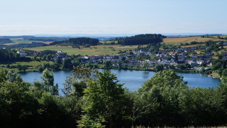 Eine ruhige Landschaft mit einem See und einem kleinen Dorf. Umgeben von grünen Bäumen und sanften Hügeln.