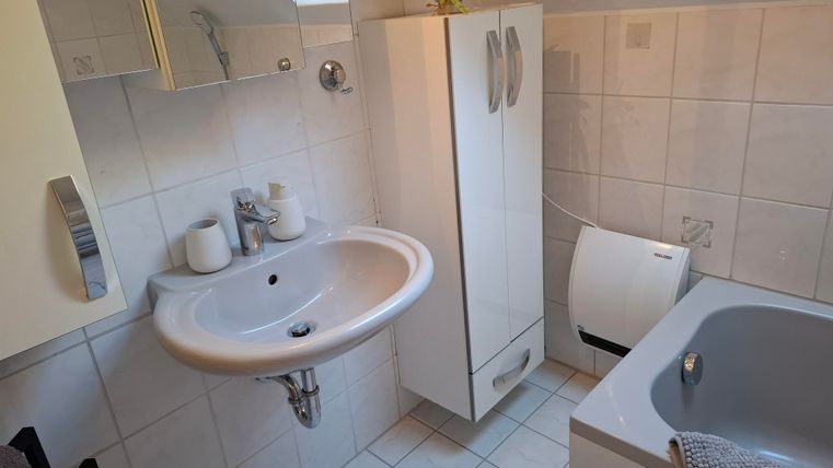 Ein modernes Badezimmer mit einem Waschbecken, einer Badewanne und einem weißen Schrank. Helle Fliesen und ein Fenster sorgen für eine angenehme Atmosphäre.