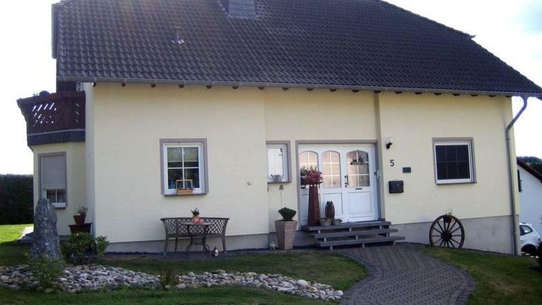 Ein gemütliches, gelbes Haus mit einem roten Dach und einem schönen Garten. Vor dem Haus stehen ein Tisch und Stühle sowie dekorative Pflanzen.