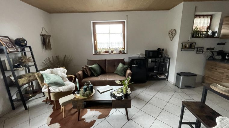 Ein gemütliches Wohnzimmer mit einer braunen Couch und einem holzbedeckten Tisch. Es gibt Pflanzen und dekorative Elemente an den Wänden.