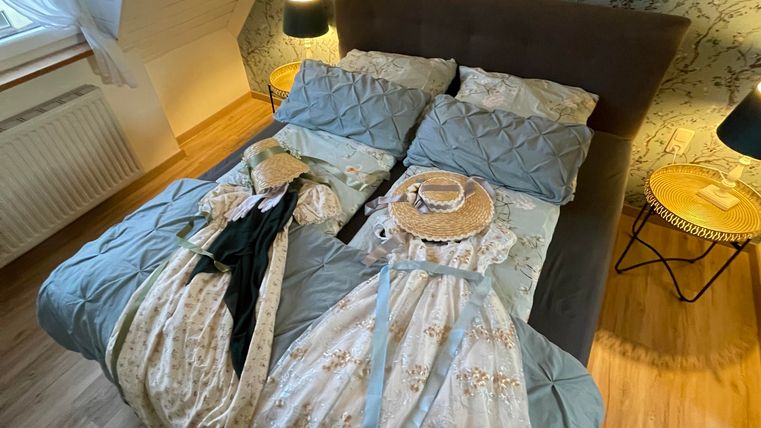 Ein Schlafzimmer mit einem großen Bett, das mit blauen und weißen Bettlaken bedeckt ist. Zwei Kleider und ein Hut liegen auf dem Bett und der Raum hat eine gemütliche Atmosphäre.