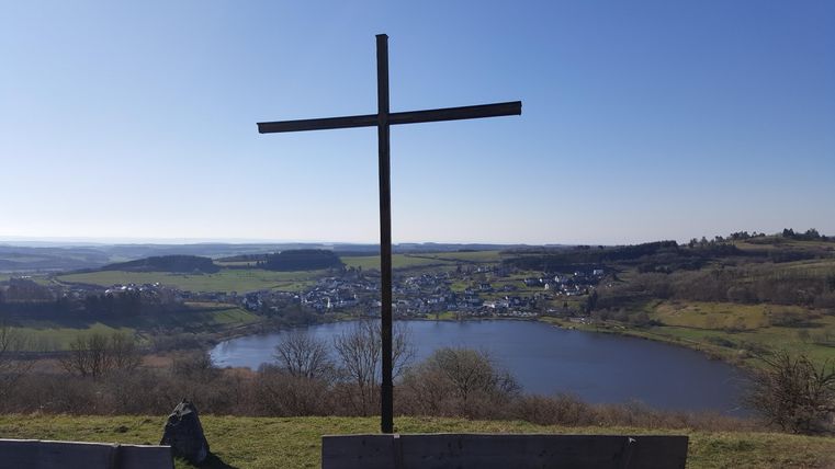 Ein großes Kreuz steht auf einem Hügel mit Blick auf einen See und umliegende grüne Landschaften. Der klare Himmel und die ruhige Szenerie vermitteln eine friedliche Atmosphäre.