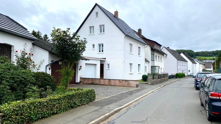 Eine ruhige Straße mit weißen Wohnhäusern und leicht bewölktem Himmel. Auf der linken Seite befinden sich kleine Gärten und parkende Autos.