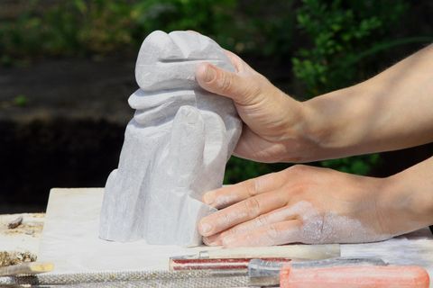 Eine Person arbeitet an einer Skulptur aus Stein. Die Hände halten einen Teil der unvollendeten Skulptur, während Werkzeuge auf dem Tisch liegen.