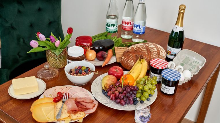 Ein Tisch mit verschiedenen Lebensmitteln, darunter Obst, Brot, Käse und Wurst. Dazu stehen Flaschen Wasser und Sekt sowie eine Blumenvase mit Tulpen.