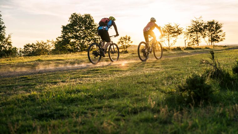 Zwei Radfahrer fahren über einen staubigen Weg bei Sonnenuntergang. Die Umgebung besteht aus grünem Gras und Bäumen im Hintergrund.