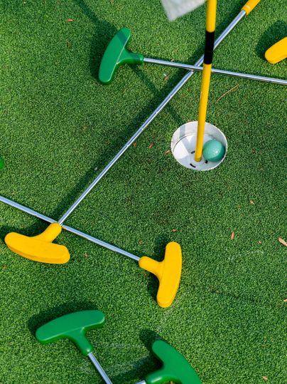 <p>In der Mitte des Bildes sieht man ein Loch, indem ein grüner Golfball liegt. Um das Loch ist grüner Kunstrasen ausgelegt, auf dem mehrere Schläger liegen. Die Schläger haben gelbe und grüne Schlägerköpfe, der Schaft ist metallfarbend.</p>