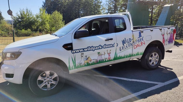 Ein weißer Pickup mit einem bunten Aufkleber, der für den Wildpark wirbt. Im Hintergrund sind Bäume und ein blauer Himmel zu sehen.