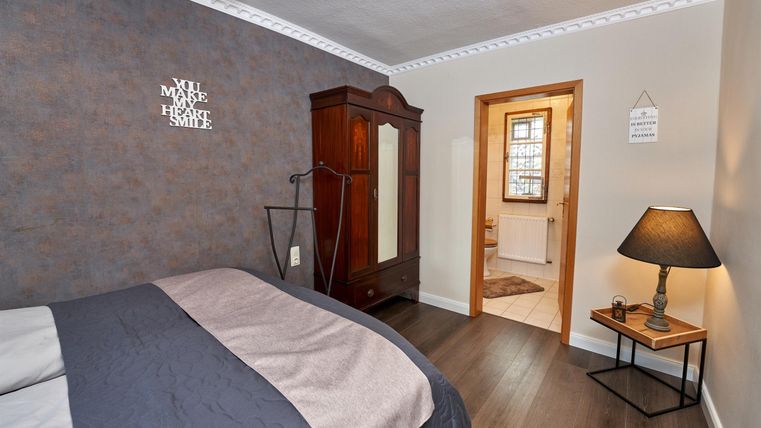 Ein gemütliches Schlafzimmer mit einem großen Bett und einem klassischen Kleiderschrank. Ein Nachttisch mit einer Lampe und ein Zugang zu einem anderen Raum sind ebenfalls sichtbar.