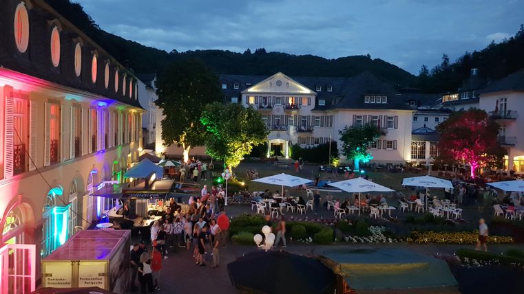 Ein lebhafter Platz bei Nacht mit bunten Lichtern und vielen Menschen. Umgeben von historischen Gebäuden und Sitzbereichen im Freien.