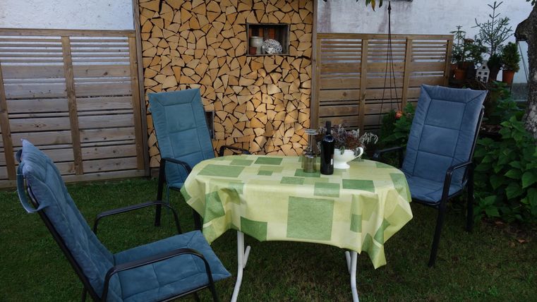 Ein gemütlicher Garten mit einem Tisch und vier Stühlen. Im Hintergrund steht ein Holzregal.