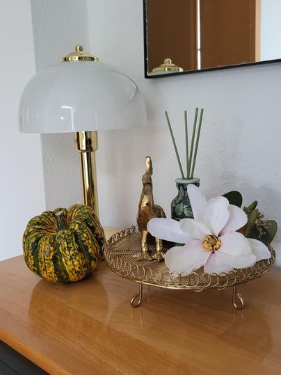 Eine dekorative Tischanordnung mit einer Lampe, einer goldenen Figur und einer Blumendekoration. Auf dem Tisch liegt ein geflochtener Korb mit einem Kürbis und anderen dekorativen Elementen.