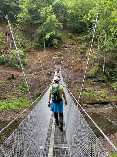 Eine Hängebrücke über einen kleinen Bach in einem grünen Wald. Eine Person geht langsam über die Brücke.