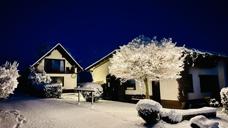 Eine schneebedeckte Landschaft bei Nacht mit zwei Häusern und einem erleuchteten Baum. Die ruhige Szene strahlt winterliche Gemütlichkeit aus.