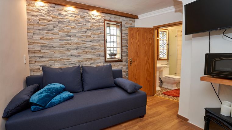 Ein gemütliches Wohnzimmer mit einer blauen Couch und Dekokissen. Im Hintergrund sind ein Fernseher und ein Zugang zum Badezimmer zu sehen.
