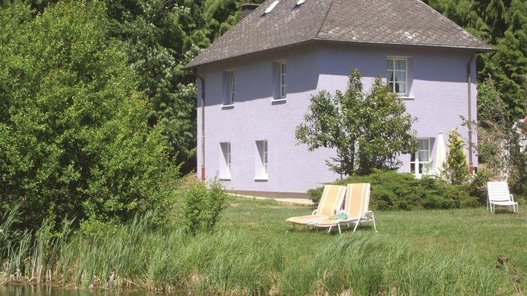 Ein gemütliches lila Haus umgeben von Bäumen und Grün. Im Vordergrund liegt ein kleiner Teich mit Liegen zum Entspannen.