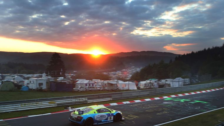 Ein Rennwagen fährt bei Sonnenuntergang auf einer berühmten Rennstrecke. Im Hintergrund sind Zeltplätze und eine malerische Landschaft zu sehen.
