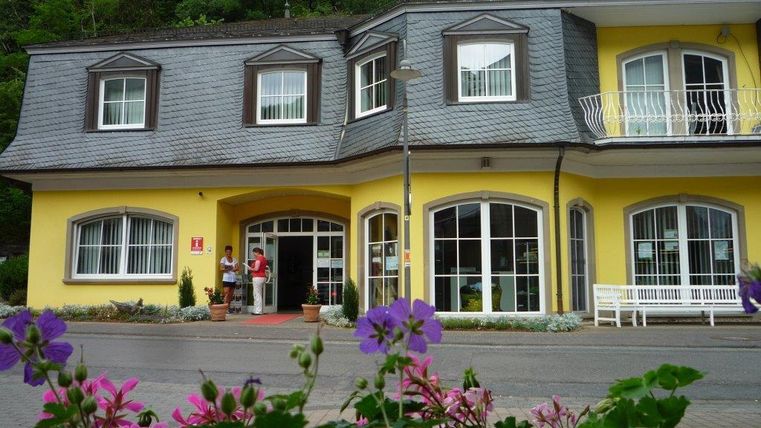 Ein gelbes Gebäude mit großen Fenstern und einem Eingangsbereich. Vor der Fassade wachsen bunte Blumen.