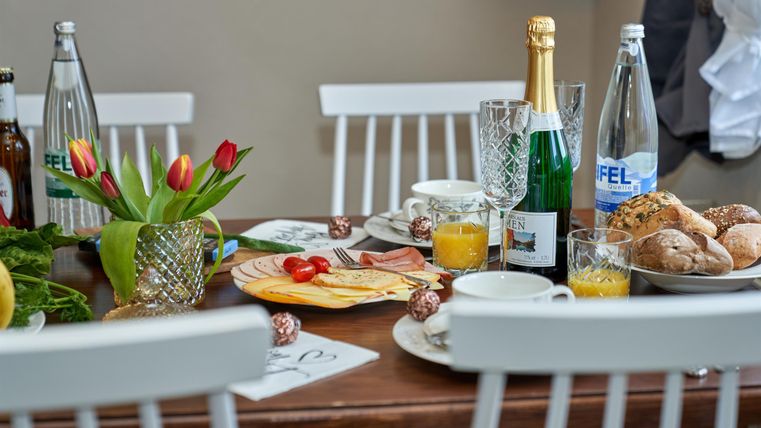 Ein schön gedeckter Tisch mit einem Frühstücksarrangement. Es gibt Säfte, Sekt, Brötchen und frische Blumen.