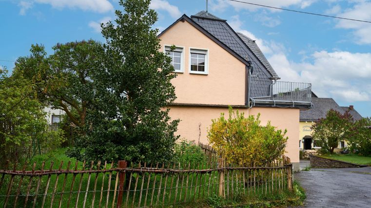 Ein hübsches Haus mit einem grauen Dach und einem kleinen Balkon. Umgeben von Garten und einem Holzzaun.
