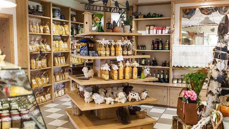 Ein gemütlicher Laden mit Holzregalen, die verschiedene Produkte wie Marmelade, Säfte und Dekorationen anbieten. Auf einem großen Tisch in der Mitte sind zusätzliche Artikel wie Plüschtiere und Pflanzen dekoriert.