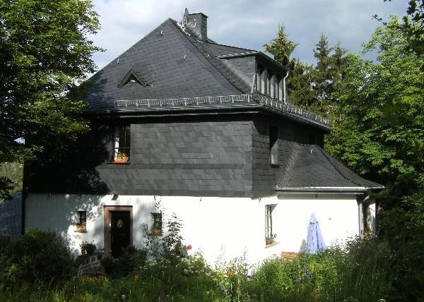 Ein charmantes Haus mit schwarzer Schindelverkleidung und einem weißen Unterbau. Es ist von Bäumen und blühenden Pflanzen umgeben.