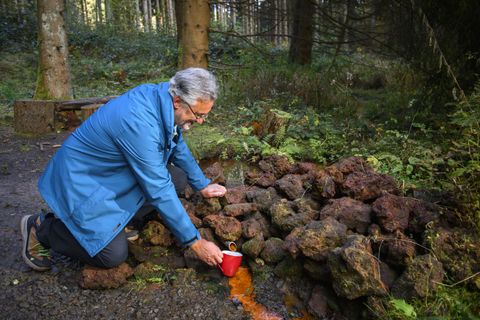 Ein Mann kniet in einem Wald und gießt eine Flüssigkeit über einen Haufen von Steinen. Die Umgebung ist grün und natürlich.