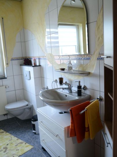Ein modernes Badezimmer mit einem Waschbecken, einem großen Spiegel und hellen Handtüchern. Die Wände sind teilweise gelb gestrichen und die Fliesen sind grau.