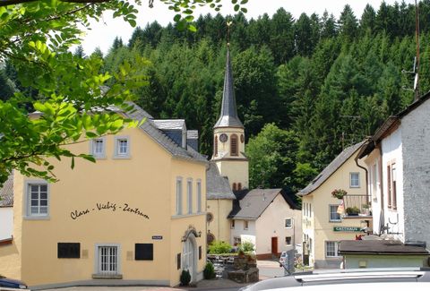 Eine malerische Dorfszene mit gelben Häusern und einem Kirchturm im Hintergrund. Die Umgebung ist von üppigem Grün umgeben.