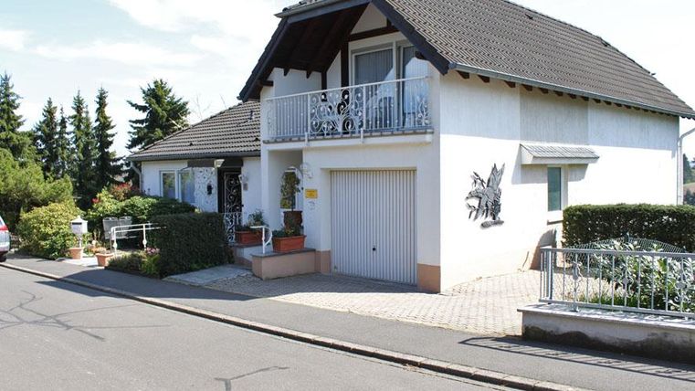A bright single-family house with a balcony and a small garden landscape. The street is quiet, surrounded by trees and plants.