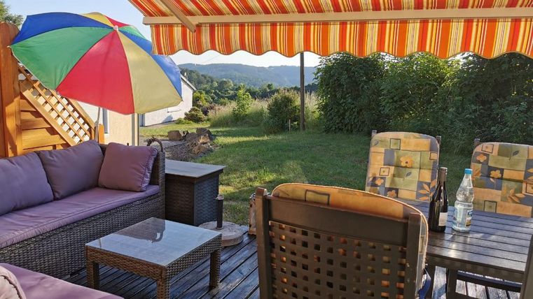Ein gemütlicher Außenbereich mit einem Sofa, einem Tisch und Stühlen. Ein bunter Sonnenschirm und eine Markise bieten Schatten, während die grüne Wiese im Hintergrund zu sehen ist.