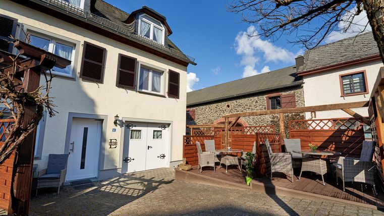 Ein gemütlicher Innenhof mit einem weißen Haus und einer Terrasse. Es gibt Gartenmöbel und einen klaren blauen Himmel.