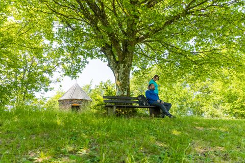 Zwei Personen sitzen lächelnd auf einer Bank im grünen unter einem Baum. Im Hintergrund befindet sich eine kleine Steinhütte.