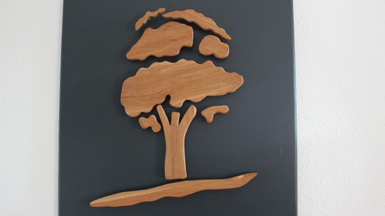 Eine Holzskulptur eines Baumes auf einer schwarzen Fläche. Der Baum hat eine stilisierte Form mit flauschigen Blättern und einem stabilen Stamm.